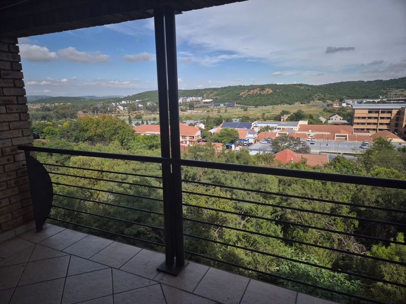 A spacious covered balcony featuring tiled flooring and a metal balustrade, offering expansive elevated views of the surrounding landscape and neighborhood.