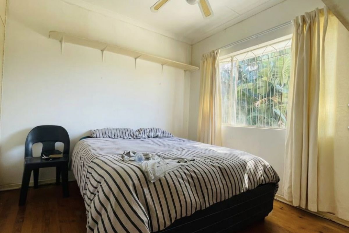 A bedroom featuring timber flooring, a ceiling fan, and a wall-mounted shelf, with natural light provided by a window with security bars.