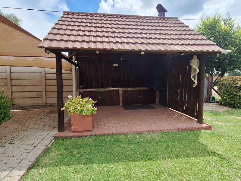 A covered outdoor patio area featuring a built-in braai, timber structure, and brick paving, set within a landscaped garden environment.