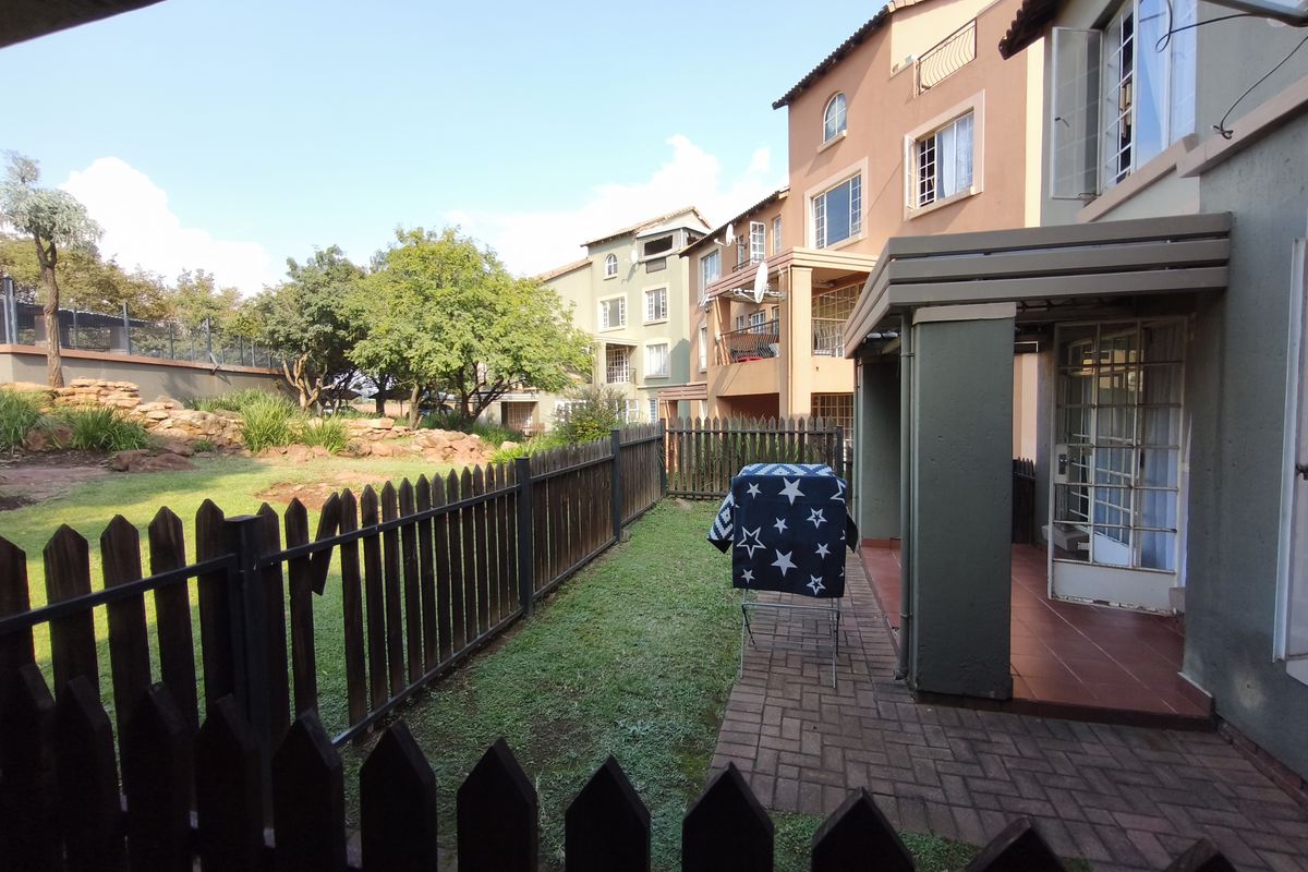 A rear view of the apartment featuring a private paved patio area, enclosed by a wooden picket fence, overlooking a communal garden space.