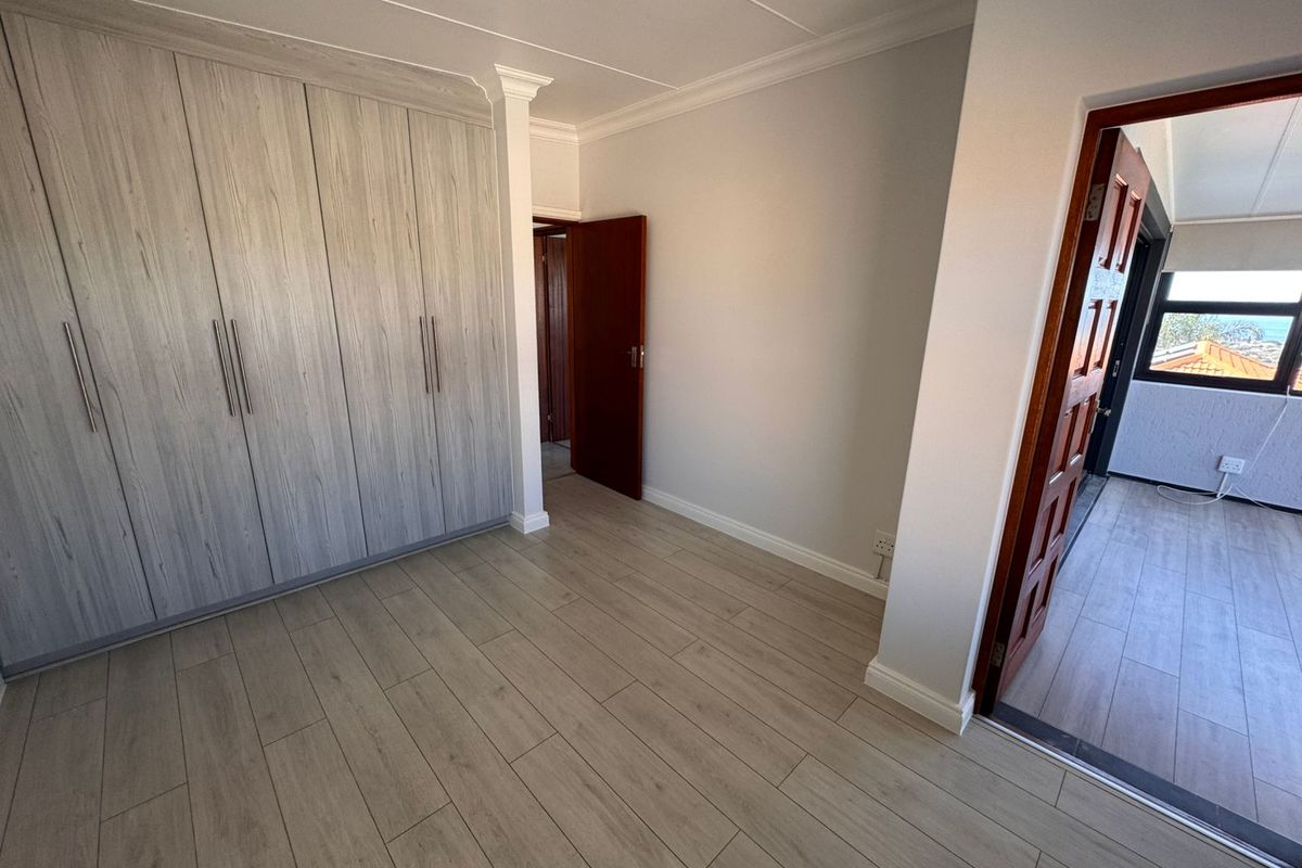 A bright, empty bedroom featuring modern light-toned laminate flooring, built-in cupboards, and classic cornicing, with an adjacent doorway leading to a balcony area.