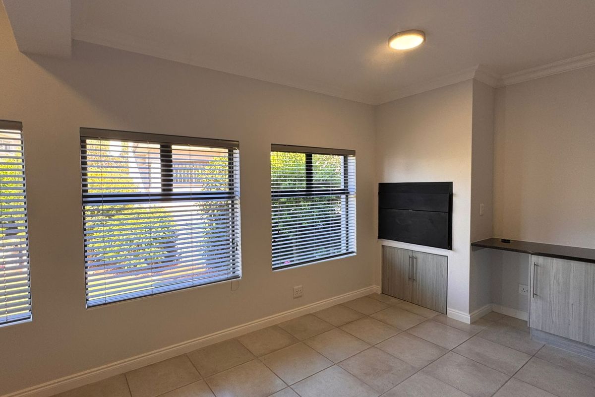 A bright, empty living area featuring neutral tiled flooring, modern built-in cabinetry, and large windows fitted with venetian blinds.