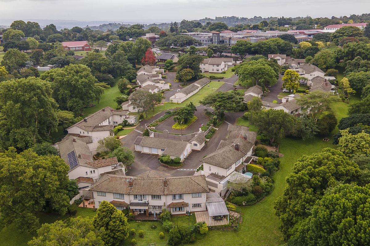 An aerial view of a well-maintained townhouse complex featuring multiple residential units, paved driveways, mature trees, and landscaped communal gardens.