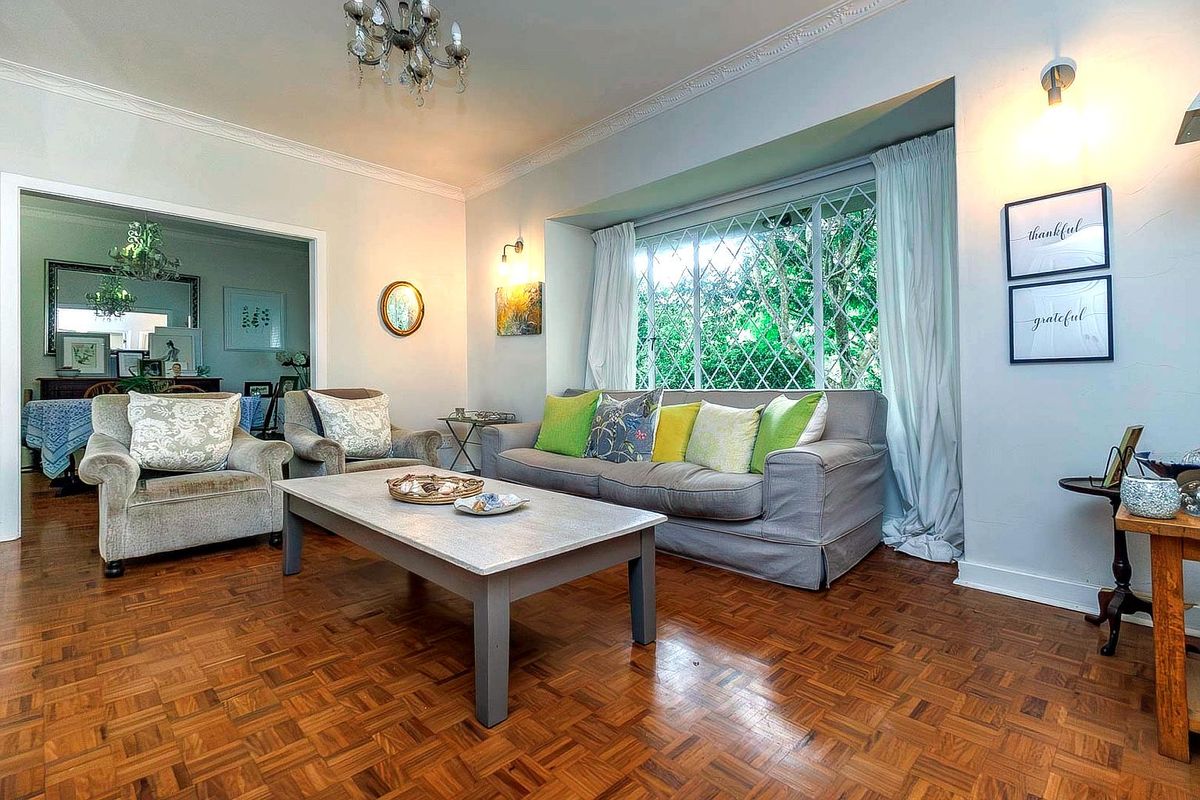A well-maintained living room featuring classic parquet flooring, decorative ceiling cornices, and a large bay window with security bars.