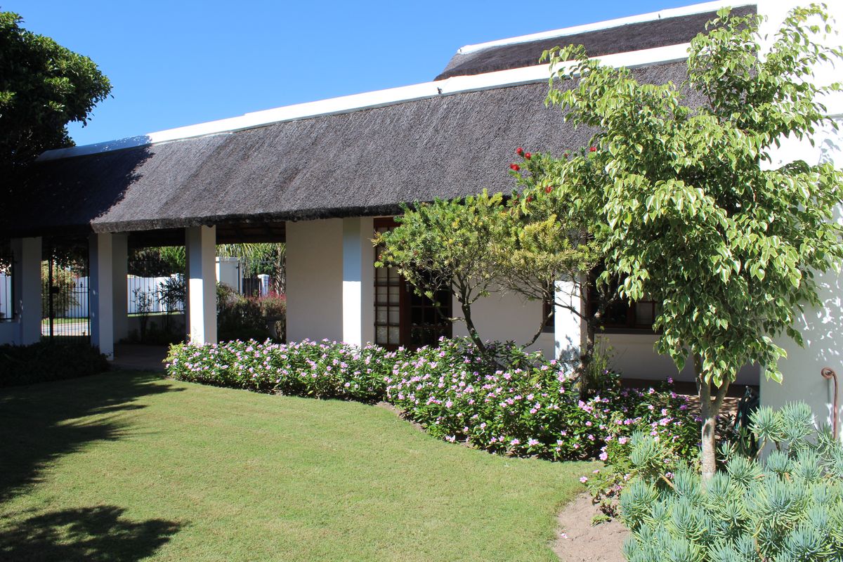 A well-maintained rear exterior view featuring a traditional thatched roof, a covered patio area, and a neatly landscaped garden with established flower beds.