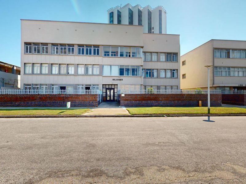 Front exterior view of a multi-story apartment building featuring a brick boundary wall, security fencing, and a central entrance.