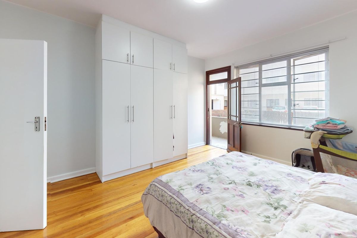 A bright bedroom featuring polished hardwood flooring, ample built-in cupboard storage, and direct access to a private balcony through a glass-paneled door.