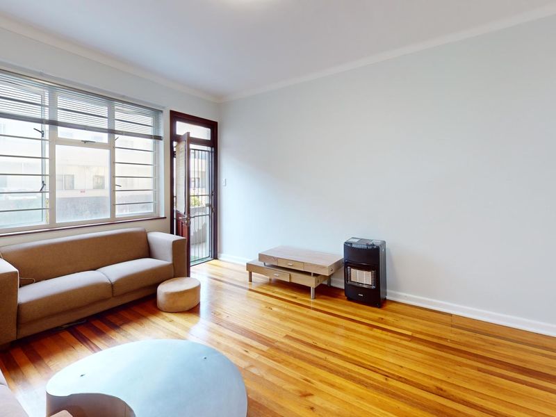 A bright living room featuring polished hardwood flooring, large windows with blinds, and a security gate leading to the exterior.