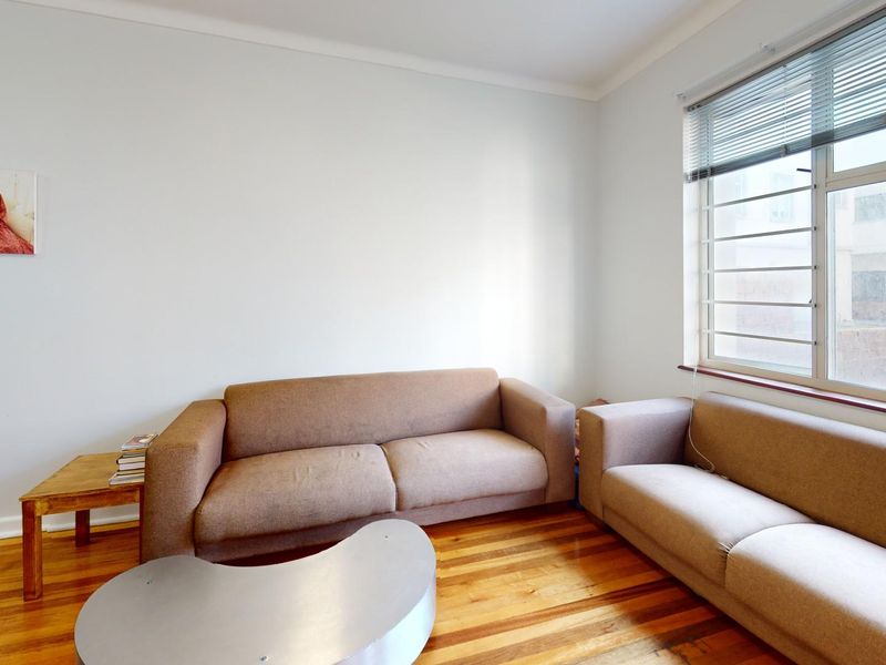A living room featuring polished timber flooring, neutral walls, and a large window with venetian blinds, providing a bright and functional living space.