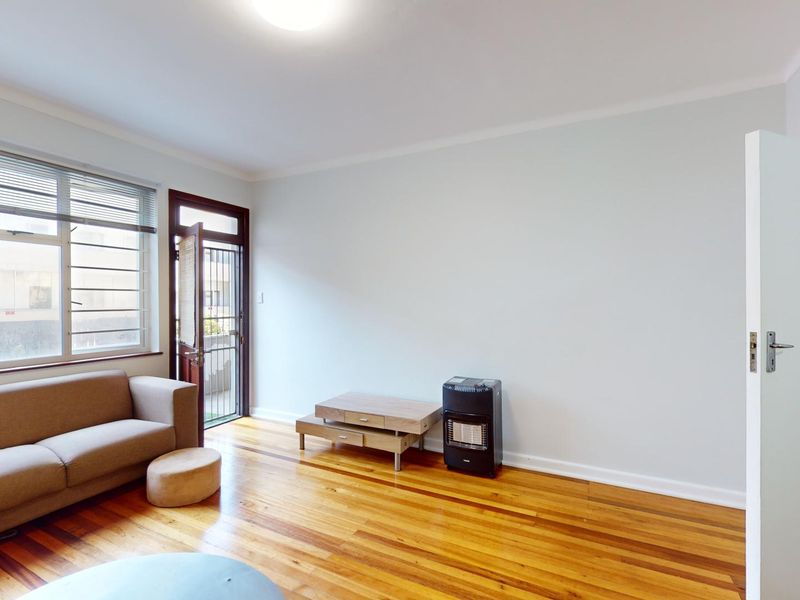 A bright living room featuring polished hardwood flooring, large windows with security bars, and direct access to an outdoor area via a security gate.
