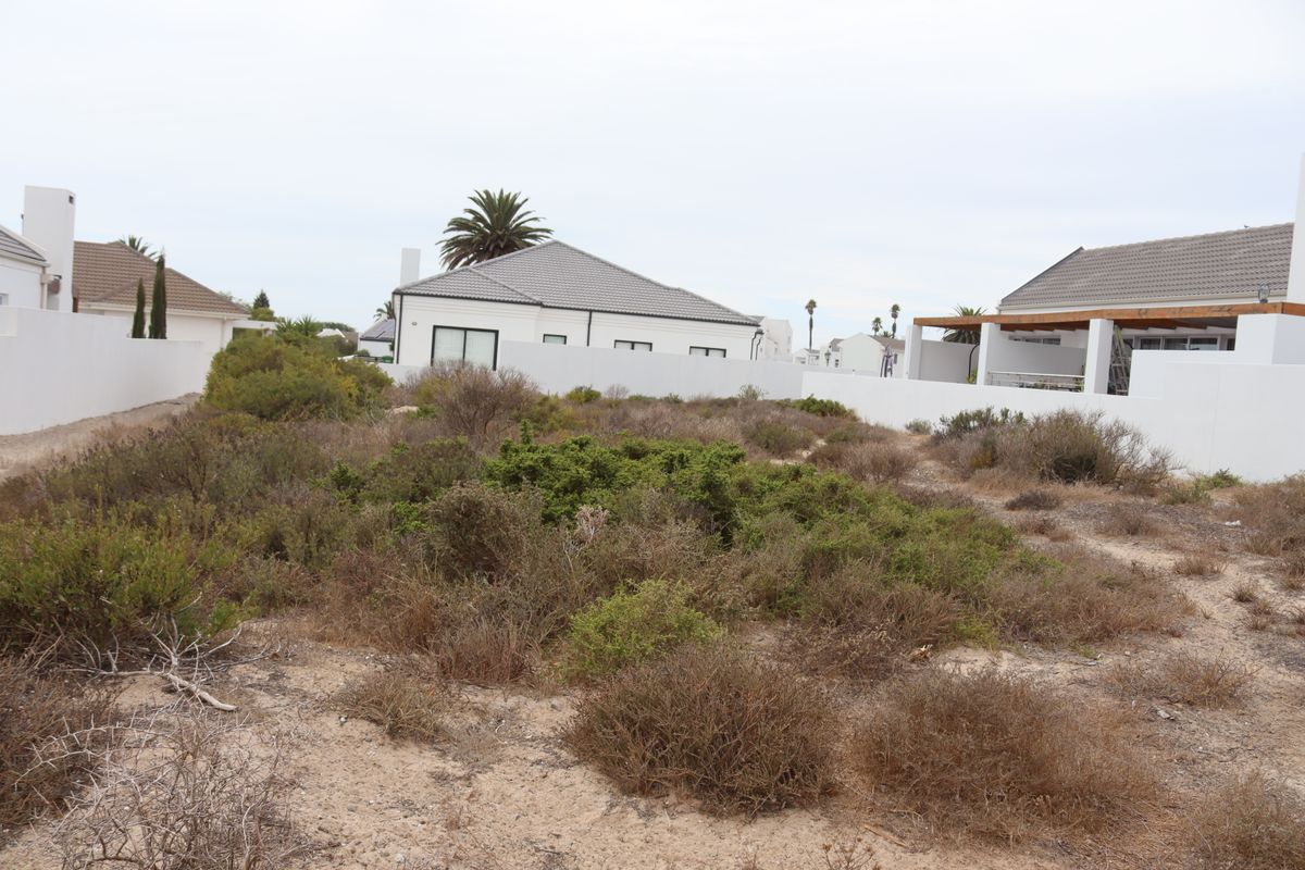 A vacant residential plot featuring natural coastal scrub vegetation, bordered by existing white boundary walls and neighbouring residential properties.