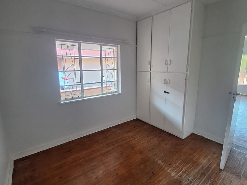 An empty bedroom featuring original timber flooring, built-in cupboards, and a window with security bars.