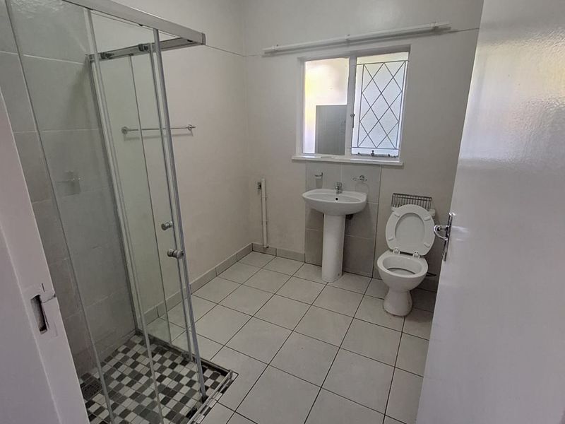 A functional bathroom featuring a glass shower enclosure, pedestal basin, and toilet with neutral tiled flooring and natural light.