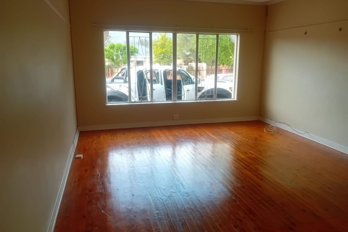 An empty living room featuring polished hardwood flooring, large windows with security bars, and neutral wall finishes.
