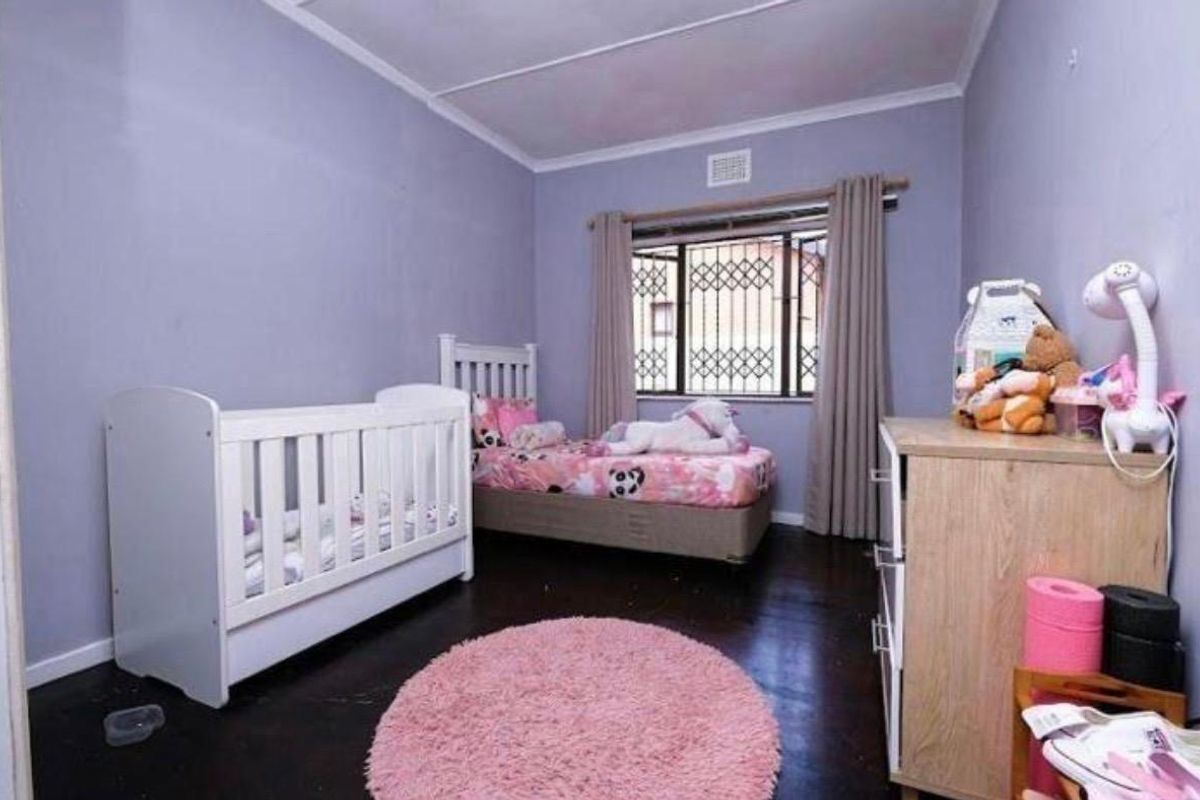 A bedroom featuring dark flooring, light purple walls, and a window fitted with a security gate.