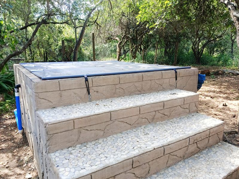 A raised outdoor pool structure featuring stone-clad steps with pebble finishes and a protective pool cover, set within a natural garden environment.