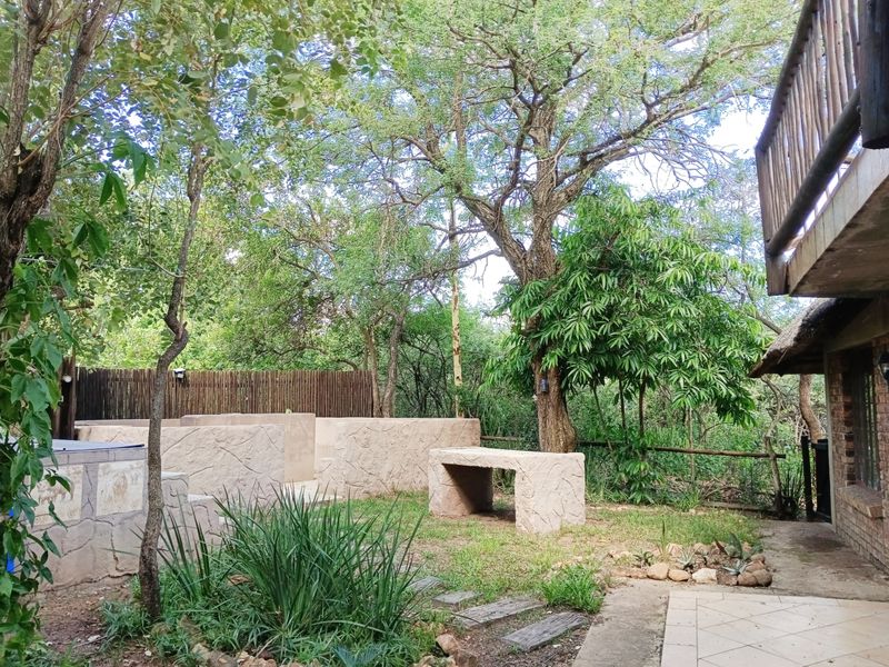 A natural garden setting featuring a built-in stone bench, retaining wall, and mature trees, adjacent to a tiled patio area.