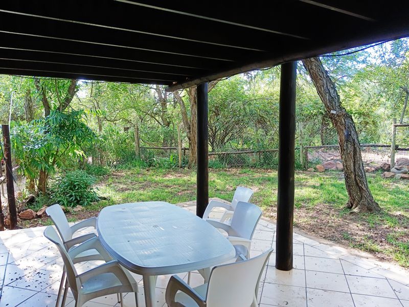 A covered outdoor patio area featuring tiled flooring and timber structural supports, overlooking a lush, tree-lined garden setting.