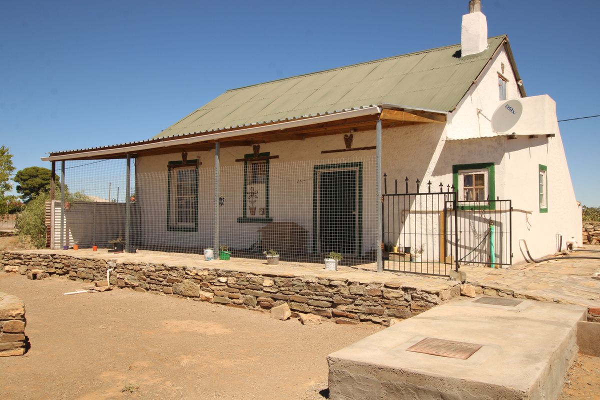 A rustic cottage featuring a stone retaining wall, covered patio area, corrugated metal roof, and a chimney, set in a rural landscape.