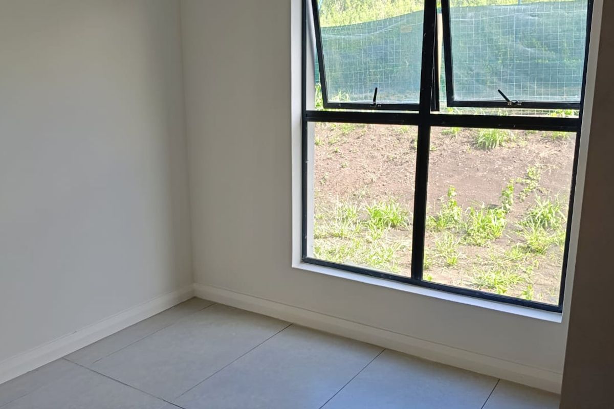 A bright, empty bedroom featuring modern grey floor tiles and a large black aluminium window frame overlooking the garden area.