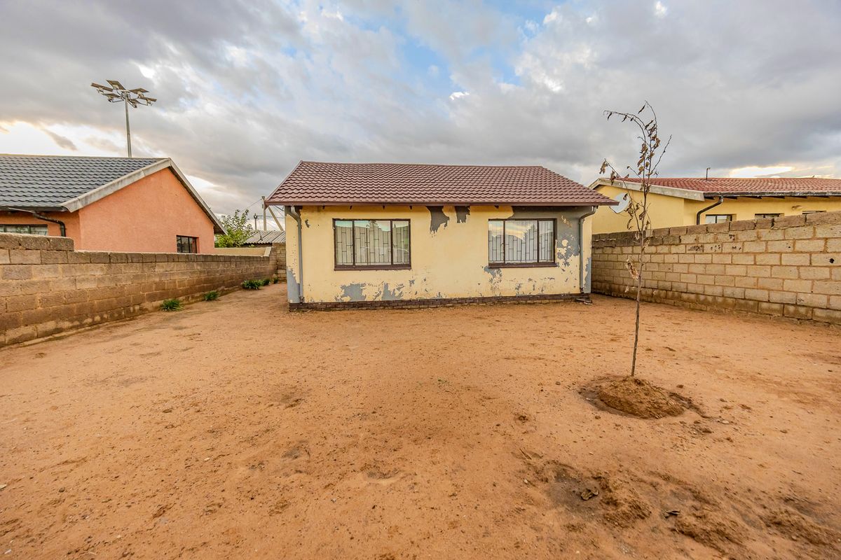 Rear view of a detached house featuring a tiled roof, security bars on windows, and a large, undeveloped sandy yard enclosed by perimeter walls.