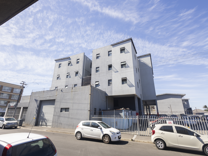 Exterior view of a multi-story apartment building featuring a grey concrete facade, secure perimeter fencing, and ground-level parking access.