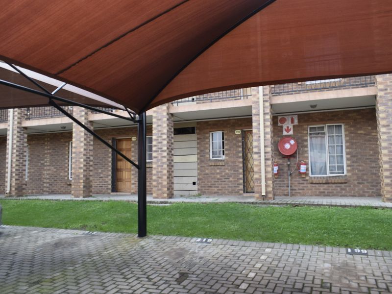 Exterior view of a townhouse complex featuring a brick facade, paved parking area with shade sail, and communal garden space.
