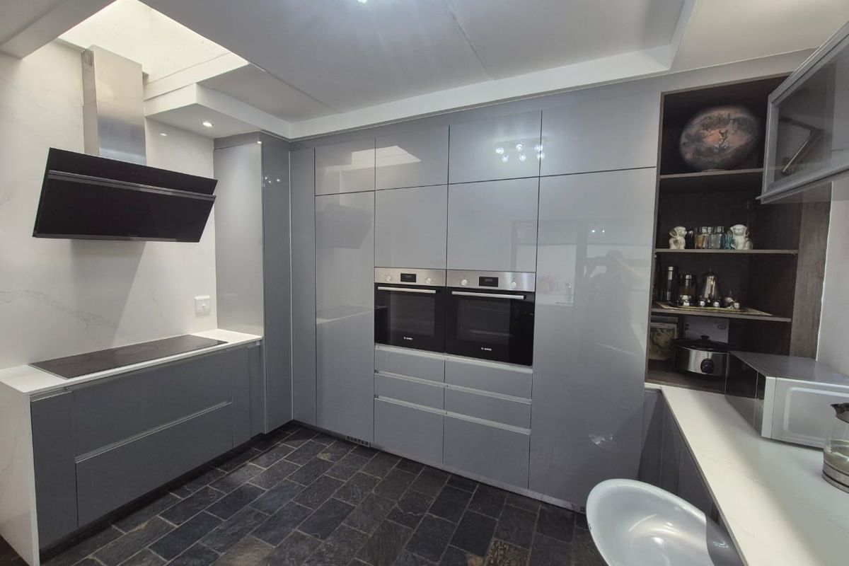 Modern kitchen featuring high-gloss grey cabinetry, integrated double ovens, a sleek extractor fan, and stone countertops with dark slate flooring.