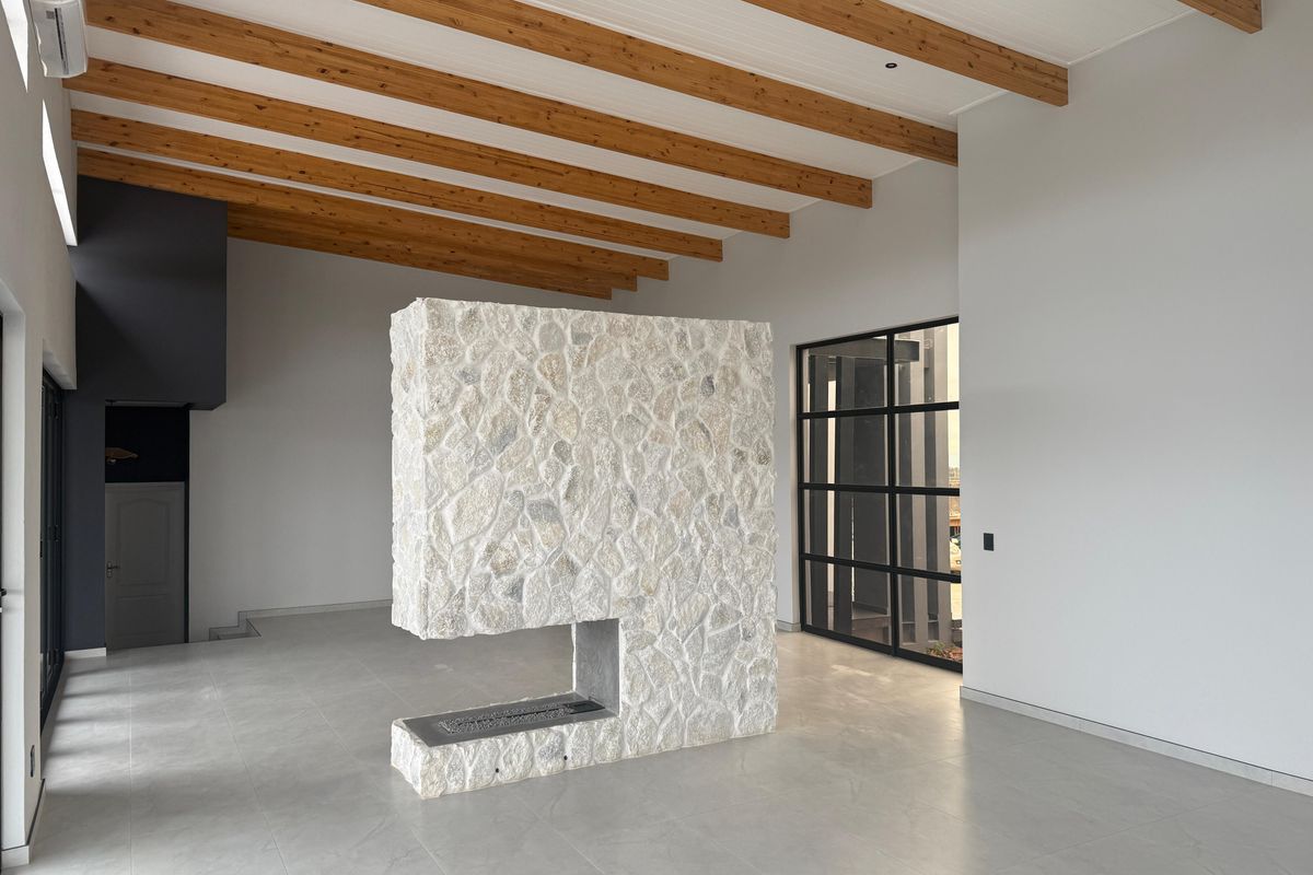 Spacious open-plan living area featuring a striking central stone fireplace, exposed timber ceiling beams, and large industrial-style black-framed windows.