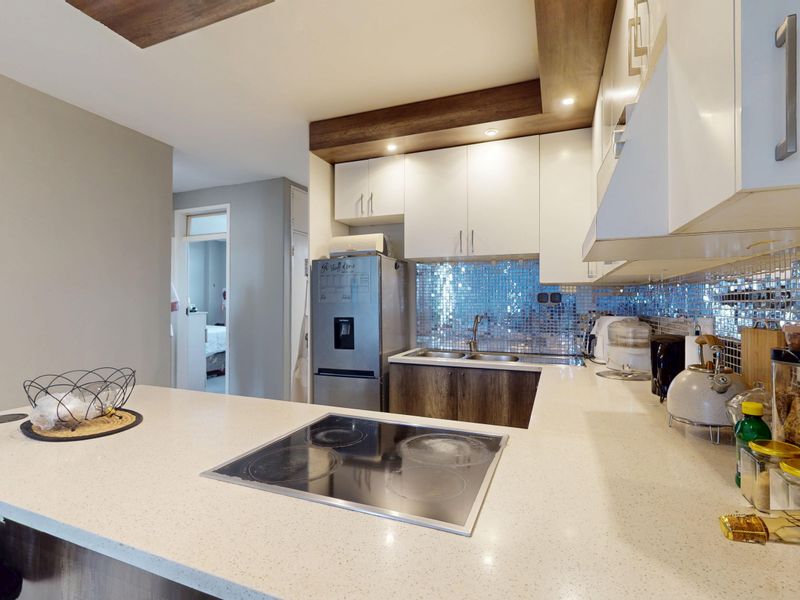 Modern kitchen featuring stone countertops, an integrated electric hob, mosaic tile backsplash, and sleek white cabinetry with wood-look ceiling accents.