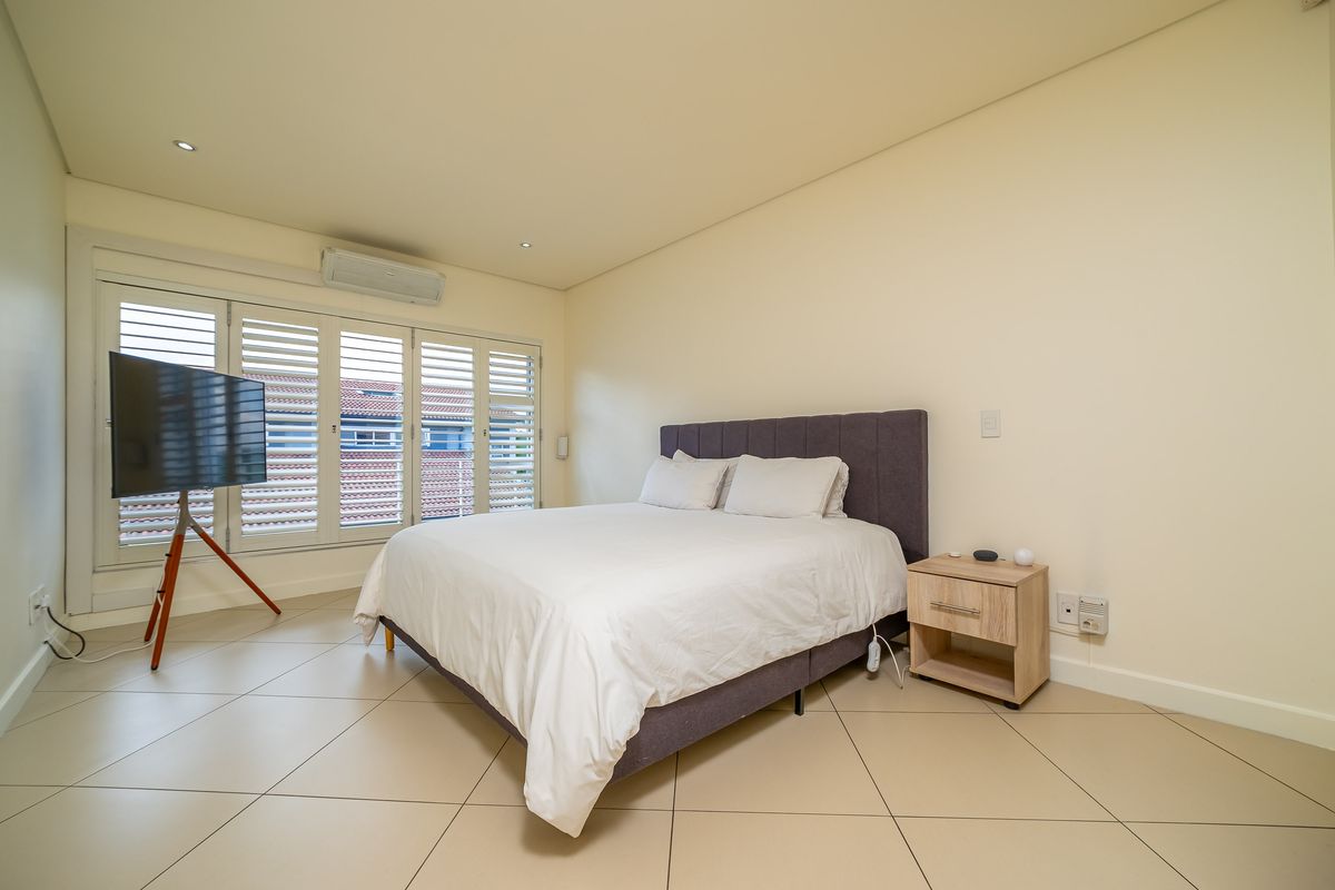 A well-maintained bedroom featuring tiled flooring, recessed lighting, a wall-mounted air conditioning unit, and white plantation shutters.