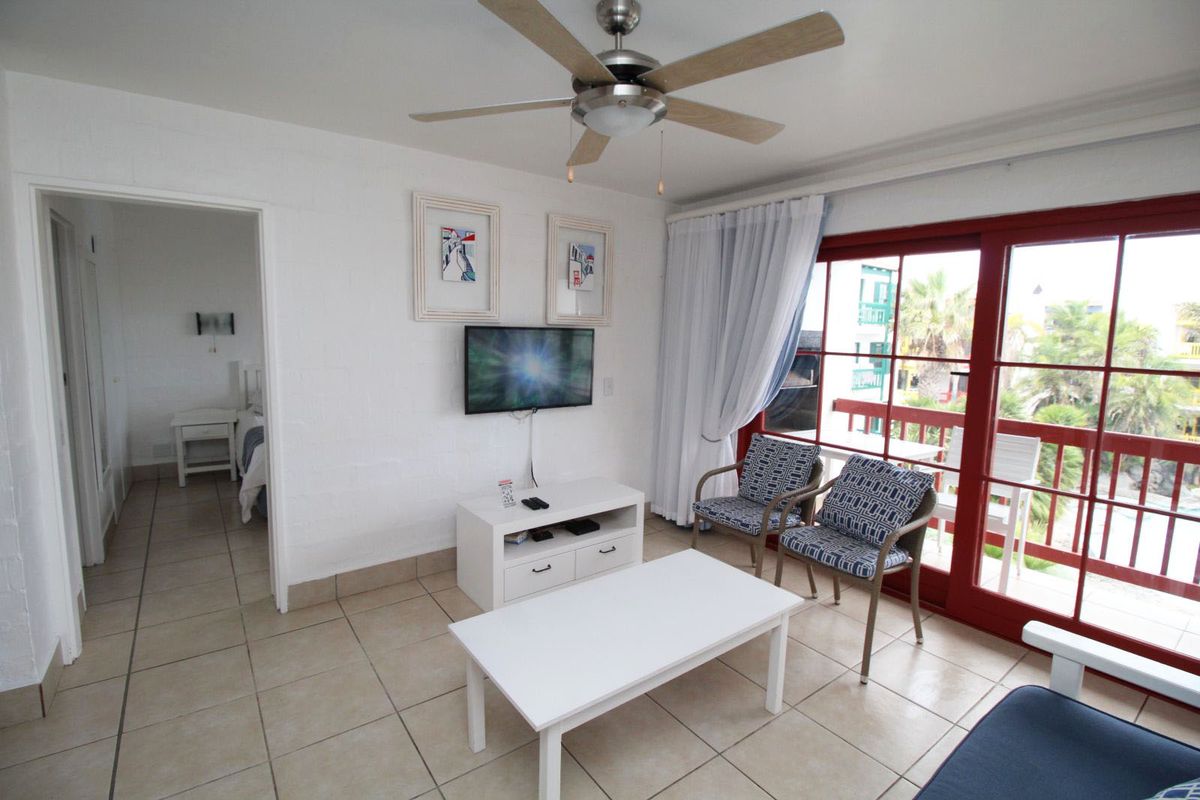 A bright living room featuring tiled flooring, a ceiling fan, and large glass doors opening onto a balcony, with an adjacent bedroom visible.
