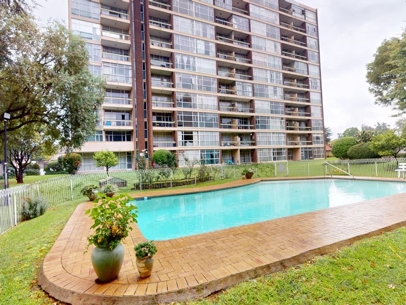 Communal swimming pool area featuring a paved surround and metal fencing, set within the landscaped grounds of a high-rise apartment complex.