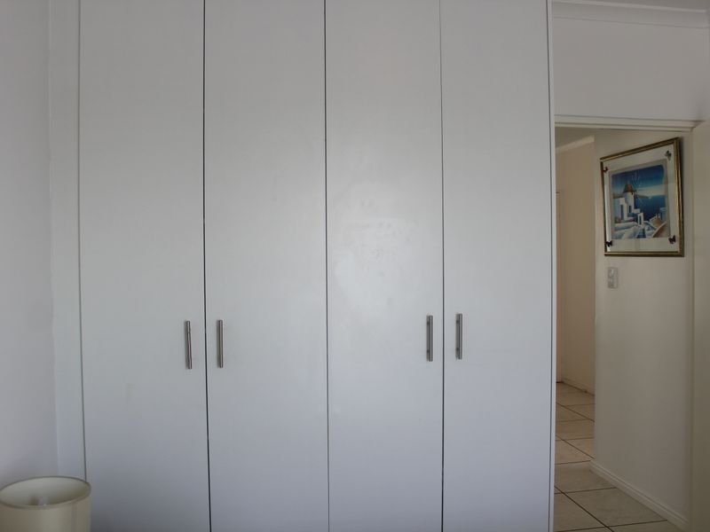 A bedroom featuring floor-to-ceiling built-in cupboards with modern handles and neutral tiled flooring, leading into an adjacent hallway.