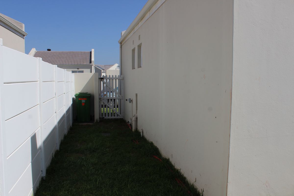 A narrow side passage of the townhouse featuring a white boundary wall, a side access gate, and a small grassed area.