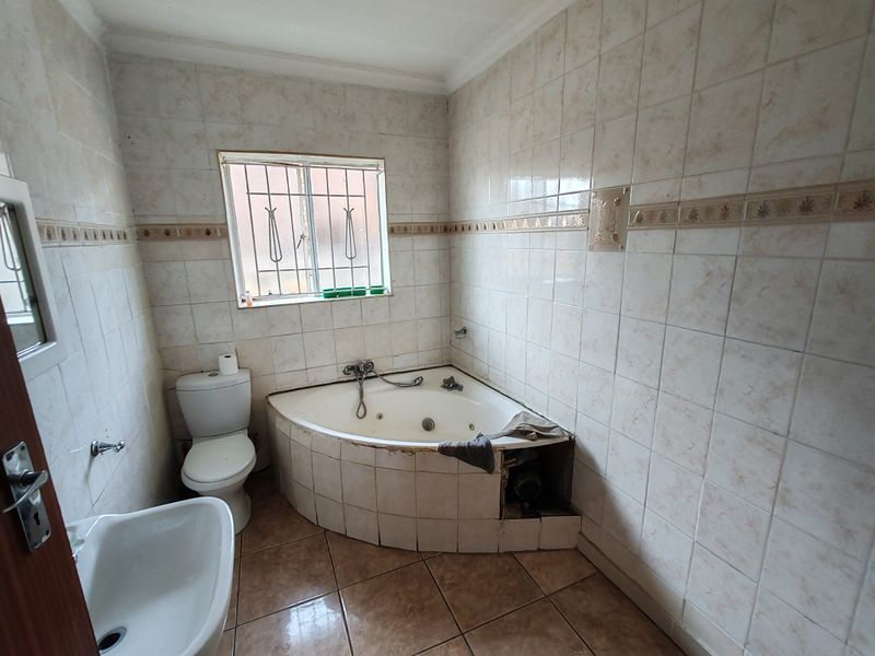 A dated bathroom featuring a corner spa bath, pedestal basin, and toilet, with full-height wall tiling and a window with security bars.