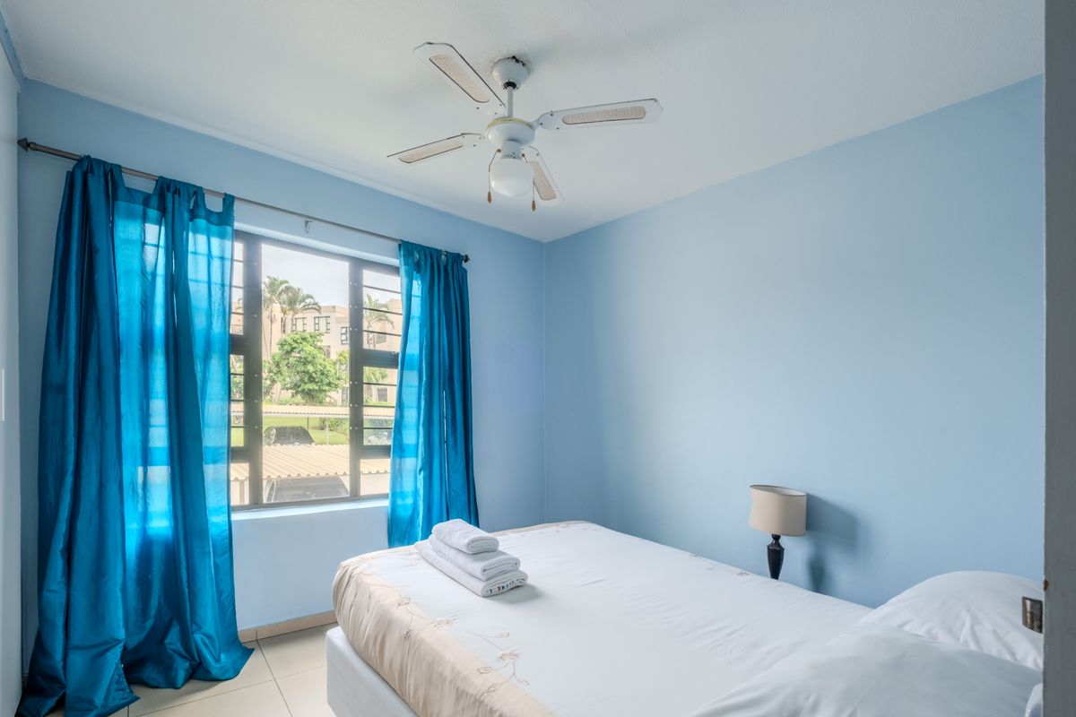 A bright bedroom featuring light blue walls, tiled flooring, a ceiling fan, and a window with security bars overlooking the complex.