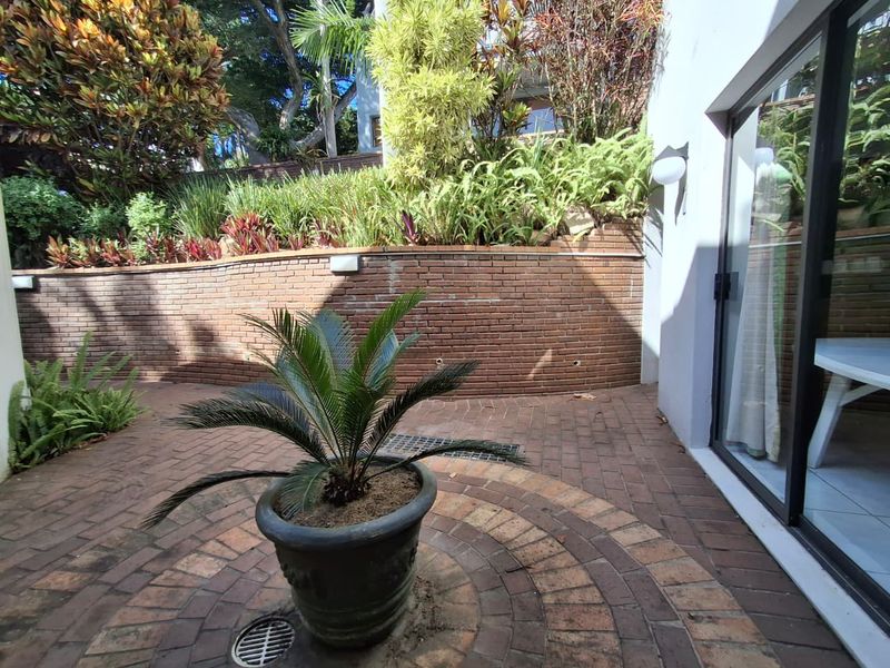 A paved outdoor patio area featuring a curved brick retaining wall, established garden beds, and direct access to the interior via sliding glass doors.