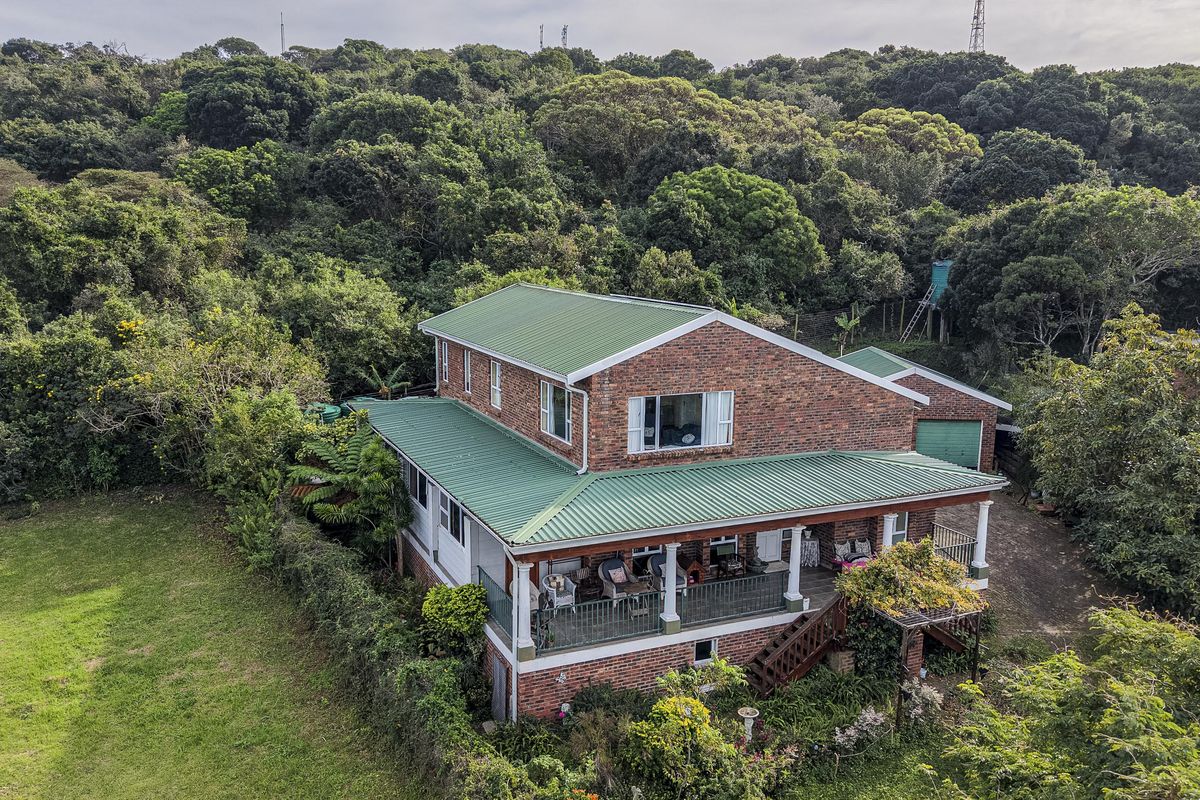Aerial view of a two-story brick house with a green metal roof, featuring a spacious covered veranda and surrounding lush garden landscape.