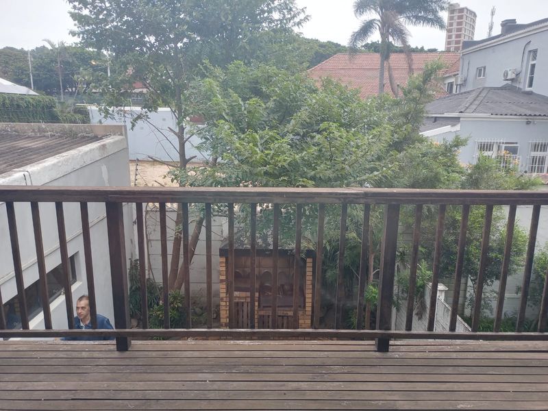 An elevated wooden balcony featuring a balustrade and a view overlooking a garden area with a built-in brick braai.