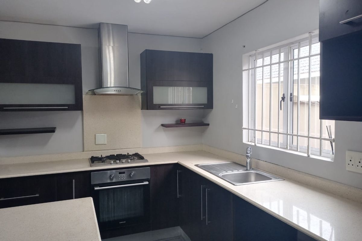 A functional kitchen featuring dark cabinetry, stone countertops, a gas hob, built-in oven, and stainless steel extractor fan with security bars on the window.