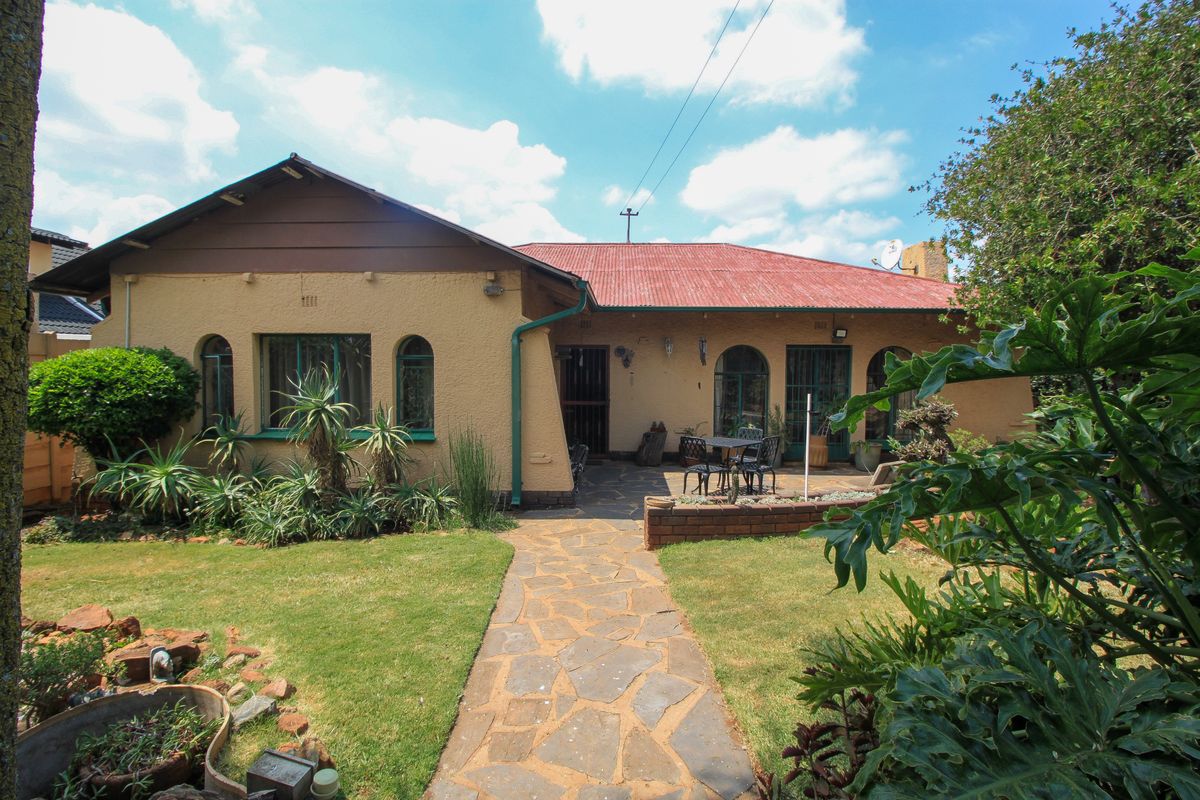 Rear view of the house featuring a stone patio, paved walkway, landscaped garden, and a corrugated metal roof.