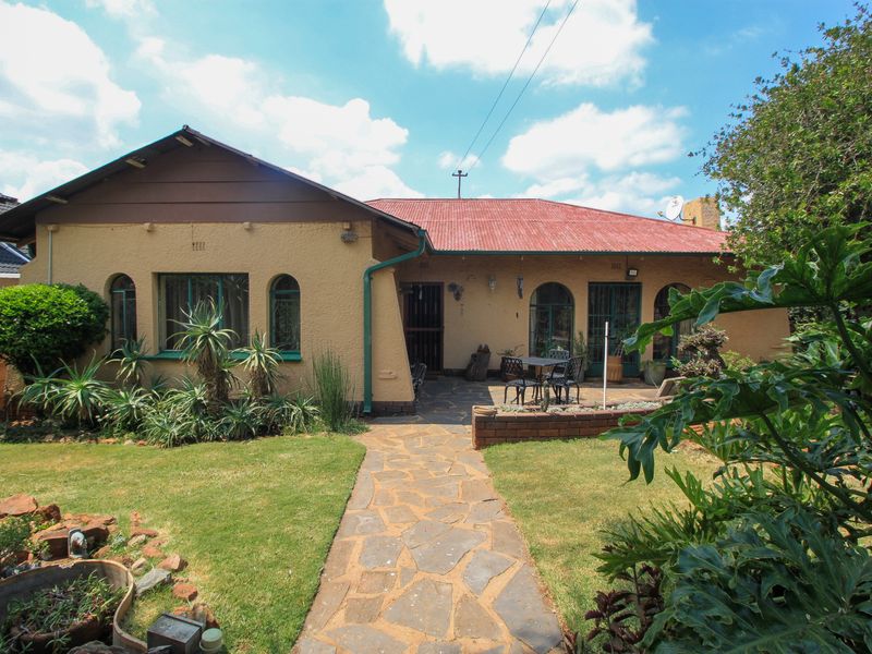 Rear view of the house featuring a stone patio, paved walkway, landscaped garden, and a corrugated metal roof.