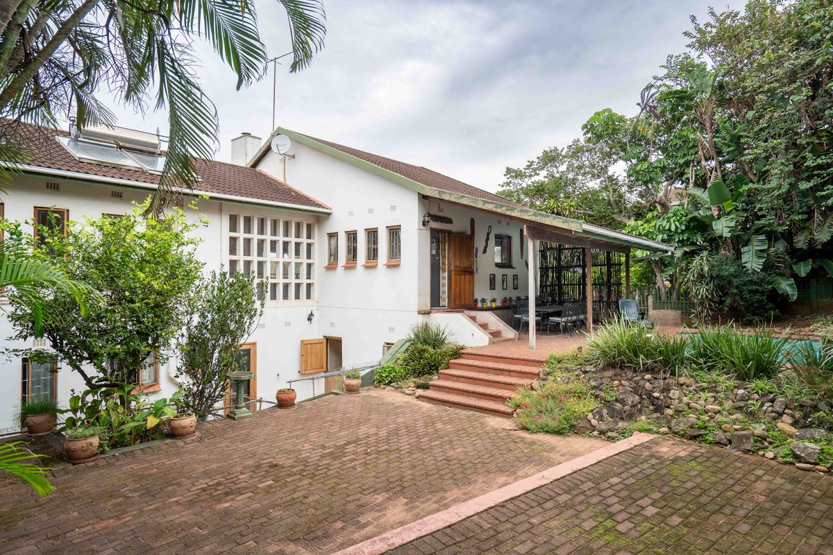 Rear view of the property featuring a brick-paved patio, covered veranda, and established garden area with stairs leading to the main dwelling.
