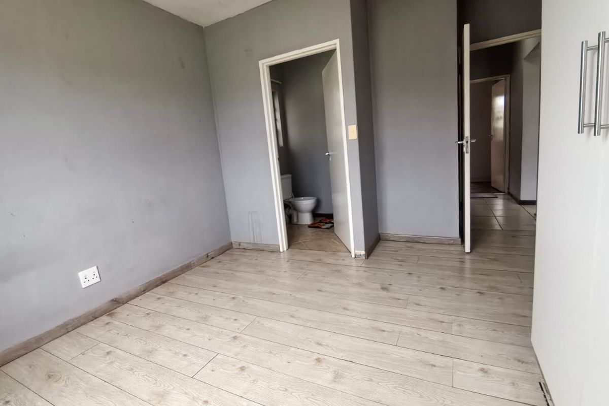 An empty bedroom featuring light-toned laminate flooring, grey painted walls, and a built-in cupboard, with an adjacent en-suite bathroom visible.