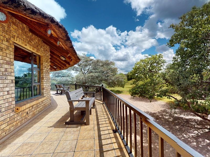 A spacious covered balcony featuring a thatched roof, face brick exterior, and metal railings, overlooking a natural garden setting.