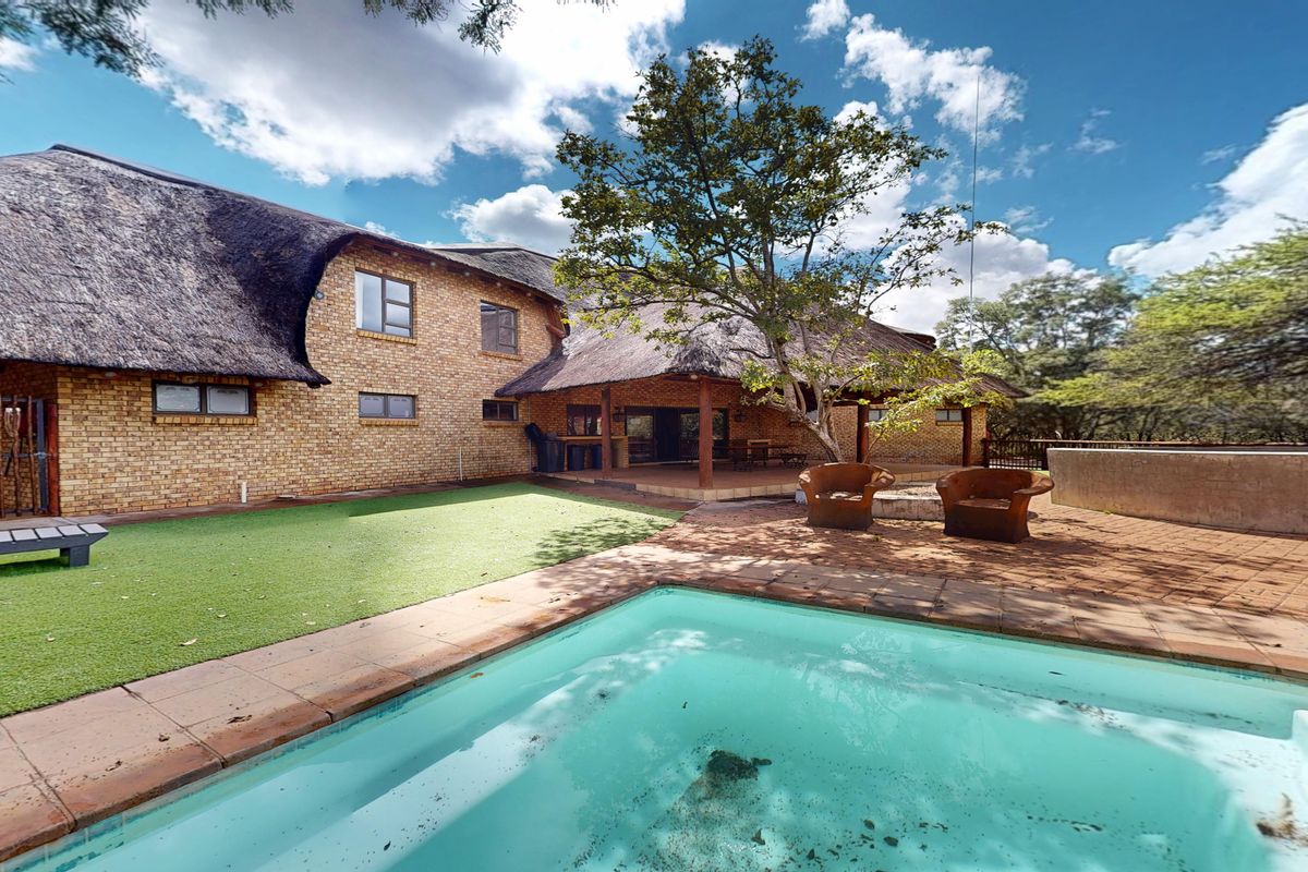 Rear view of a brick house with a thatched roof, featuring a swimming pool, covered patio area, and landscaped garden space.