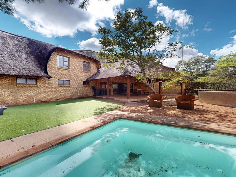 Rear view of a brick house with a thatched roof, featuring a swimming pool, covered patio area, and landscaped garden space.