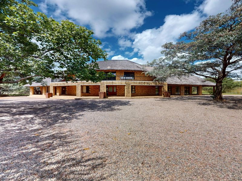 A spacious single-story house with a traditional brick facade, thatch roof, and a large gravel driveway set in a natural landscape.