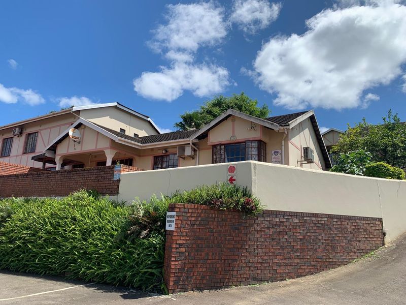 Front exterior view of a townhouse complex featuring a brick boundary wall, tiled roof, and established garden landscaping under a clear blue sky.